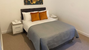 Un lit bien fait, recouvert d’une couverture grise, agrémenté d’un coussin marron et d’un coussin blanc. Une table de chevet, sur laquelle se trouvent un téléphone et une serviette pliée, est placée à côté du lit. Un tableau est accroché au-dessus du lit.