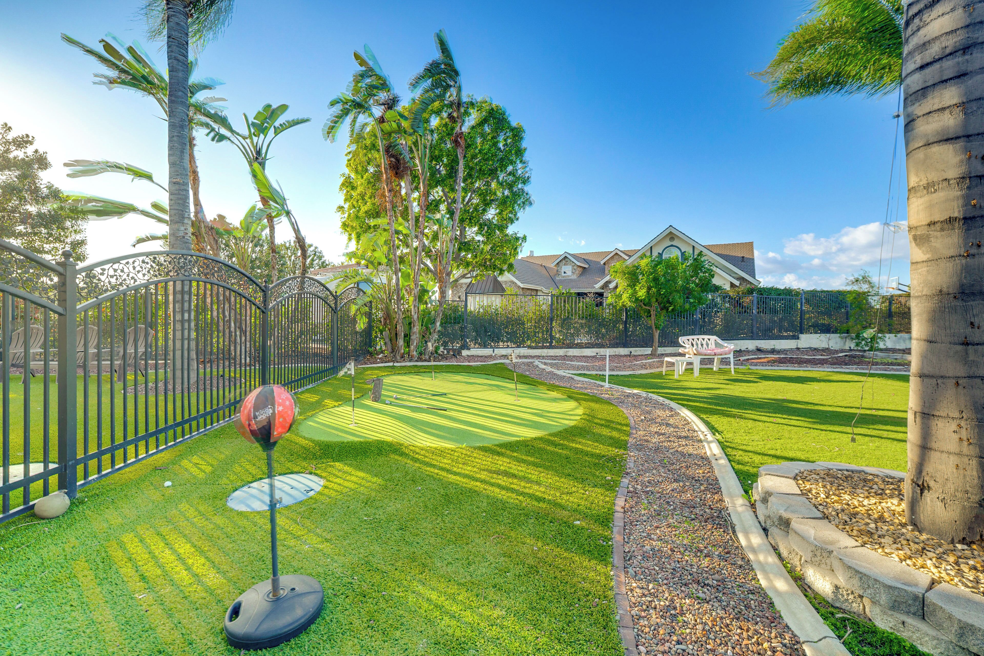 A well-maintained golf course with a putting green, surrounded by a metal fence, palm trees, and a house in the background.