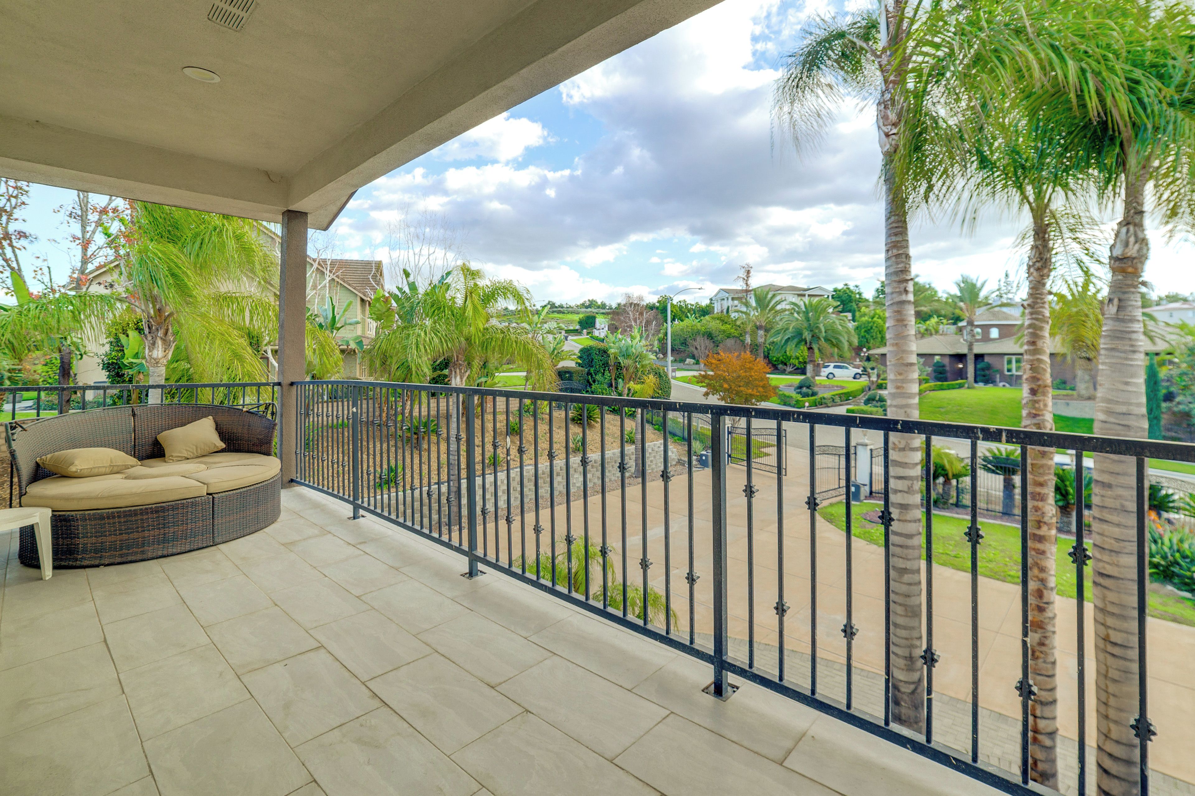 Un balcón techado con un sofá de mimbre, barandilla y vistas a una zona residencial con palmeras y casas.