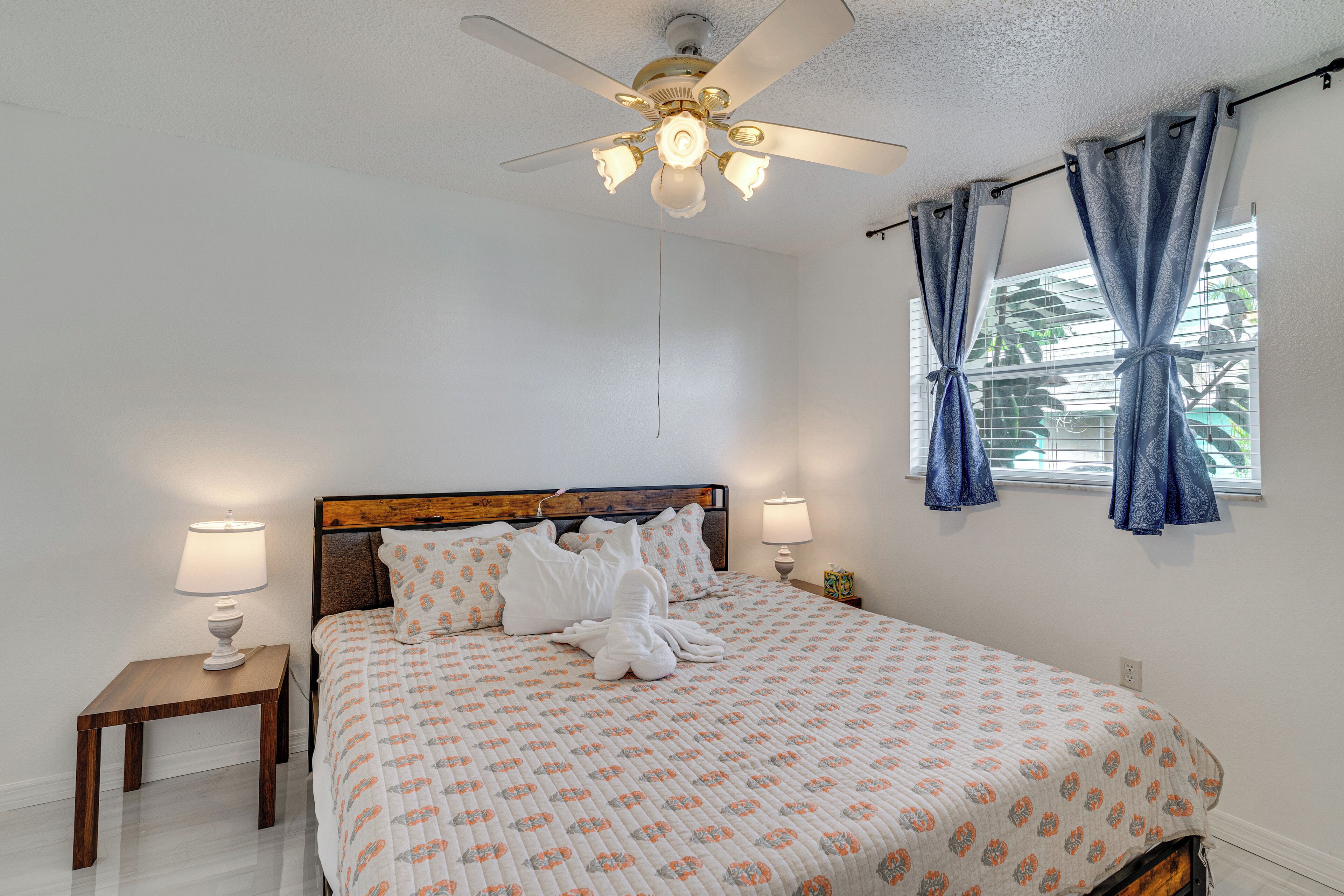 A bedroom with a wooden headboard, a bed with a floral patterned bedspread, two bedside lamps, a ceiling fan, and a window with curtains.