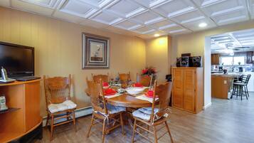 A dining area with a wooden table and chairs, a television on a stand, and a kitchen in the background.