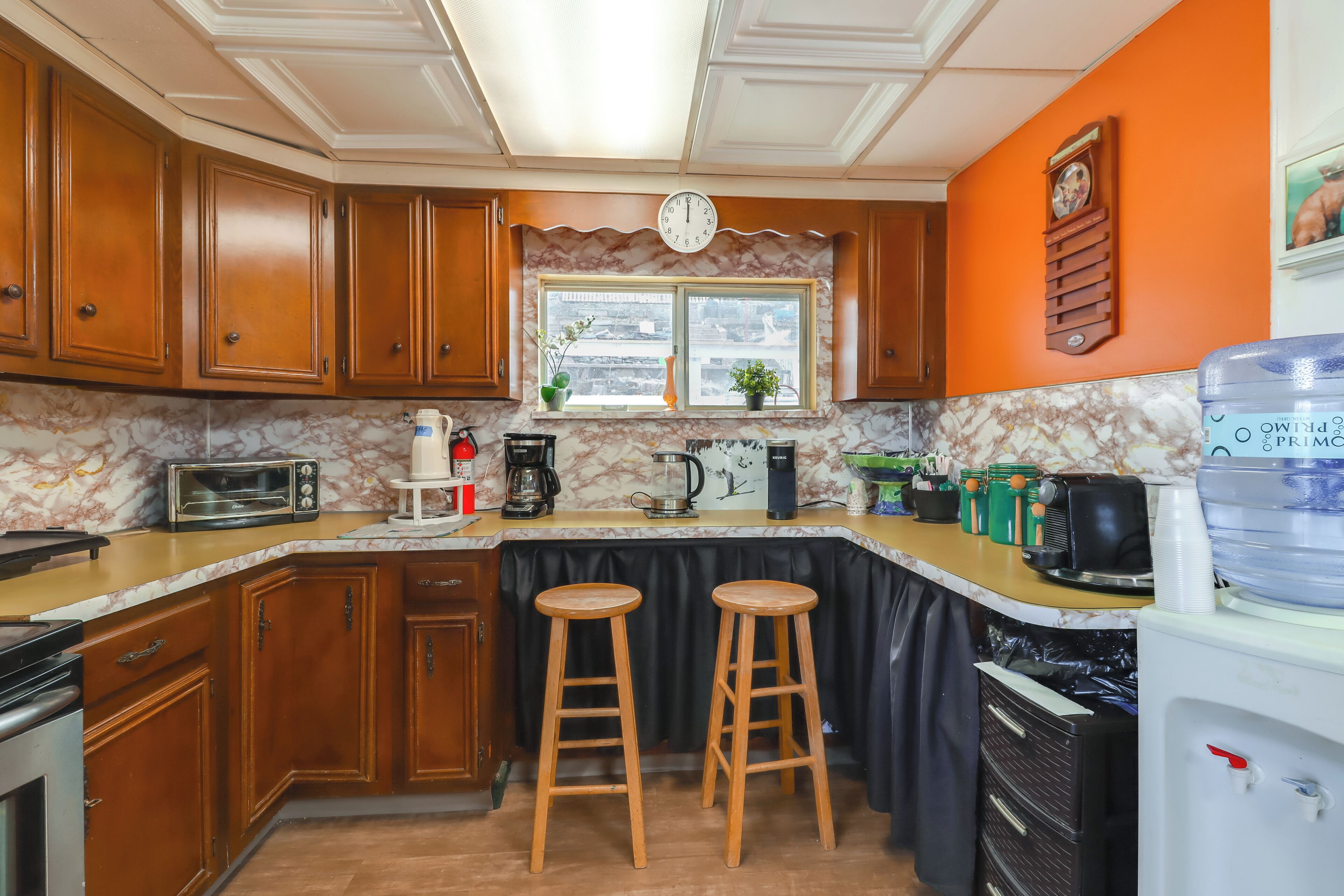 Una cocina con armarios de madera, encimera de mármol, pileta, taburetes, cafetera y refrigerador.