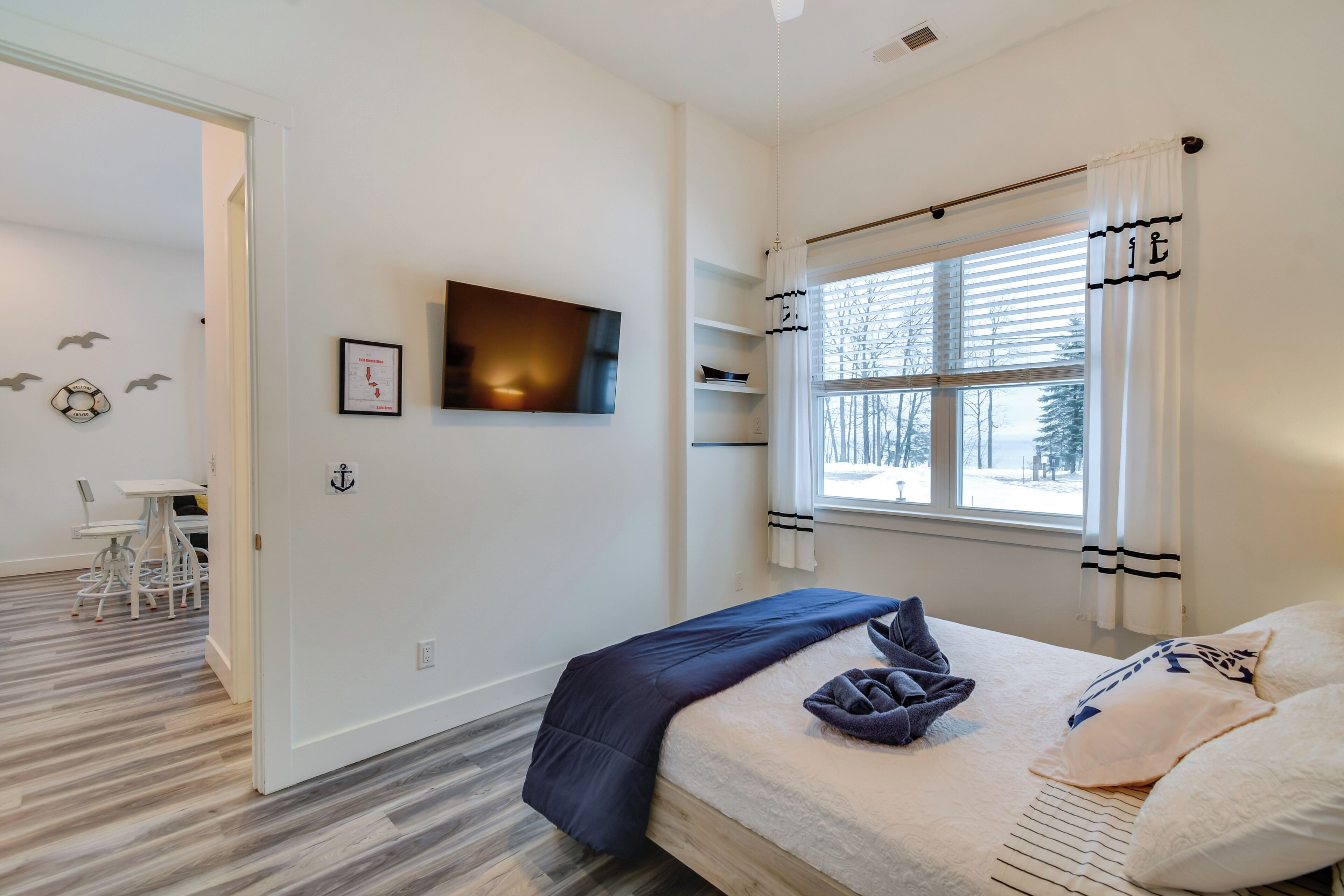A bedroom with a bed, a TV, a window with curtains, a desk with chairs, and a wall with decorative elements.