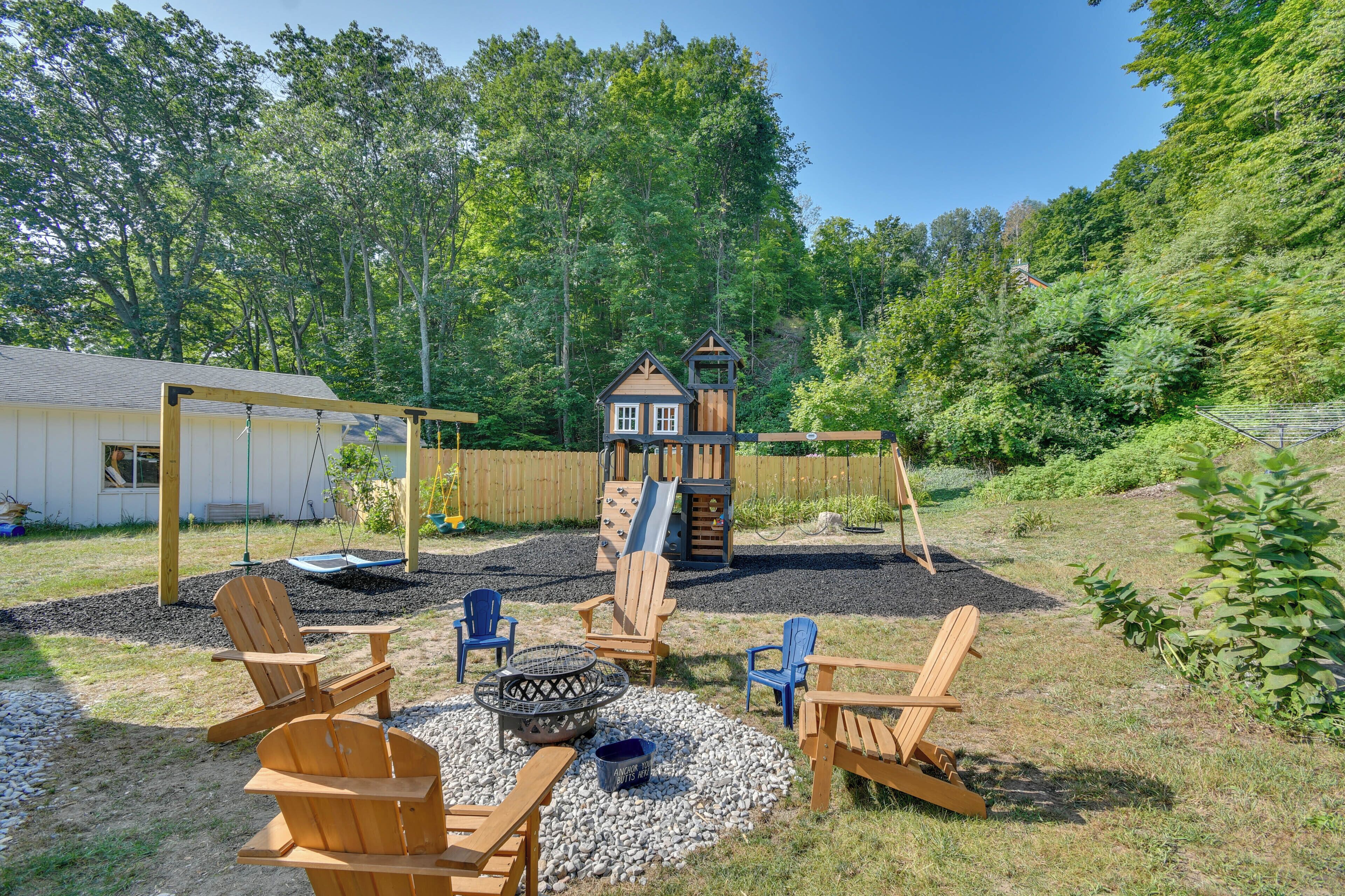 A backyard with a wooden playhouse, a swing set, Adirondack chairs, and a fire pit.