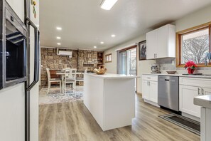 A modern kitchen with a central island, white cabinetry, a stone accent wall, and a view of the outdoors through a window. - 2 Mi to Golf Course! Family Home in Pueblo West (Pueblo)
