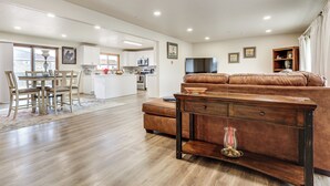 A spacious living area with a brown leather sofa, wooden coffee table, and a dining table with chairs. A kitchen is visible in the background. - 2 Mi to Golf Course! Family Home in Pueblo West (Pueblo)