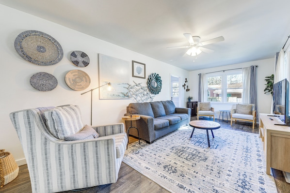 A living room with a grey sofa, striped armchair, and a round coffee table. There are wall art pieces and a ceiling fan. - 1 Mi to Park Circle: Pet-friendly Home w/ Yard (North Charleston)