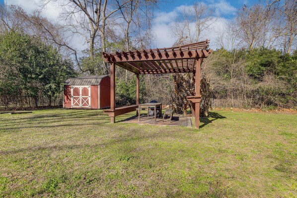 Una pérgola de madera con una mesa y sillas debajo, un cobertizo pequeño y un área con césped y árboles al fondo.