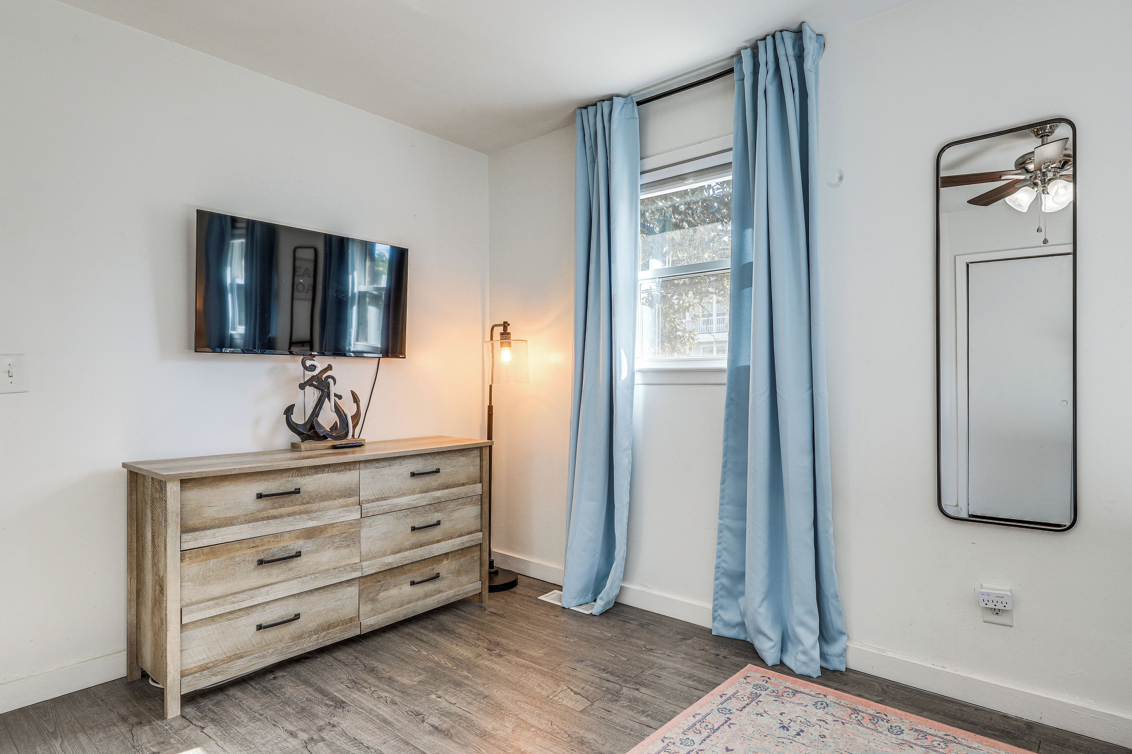 A room with a wooden dresser, a flat-screen TV mounted on the wall, a ceiling fan, and a window with blue curtains.
