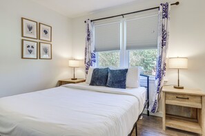 Ein ordentlich bezogenes Bett mit weißer Bettwäsche, zwei Nachttische mit Lampen und ein Fenster mit blau-weißen Vorhängen.