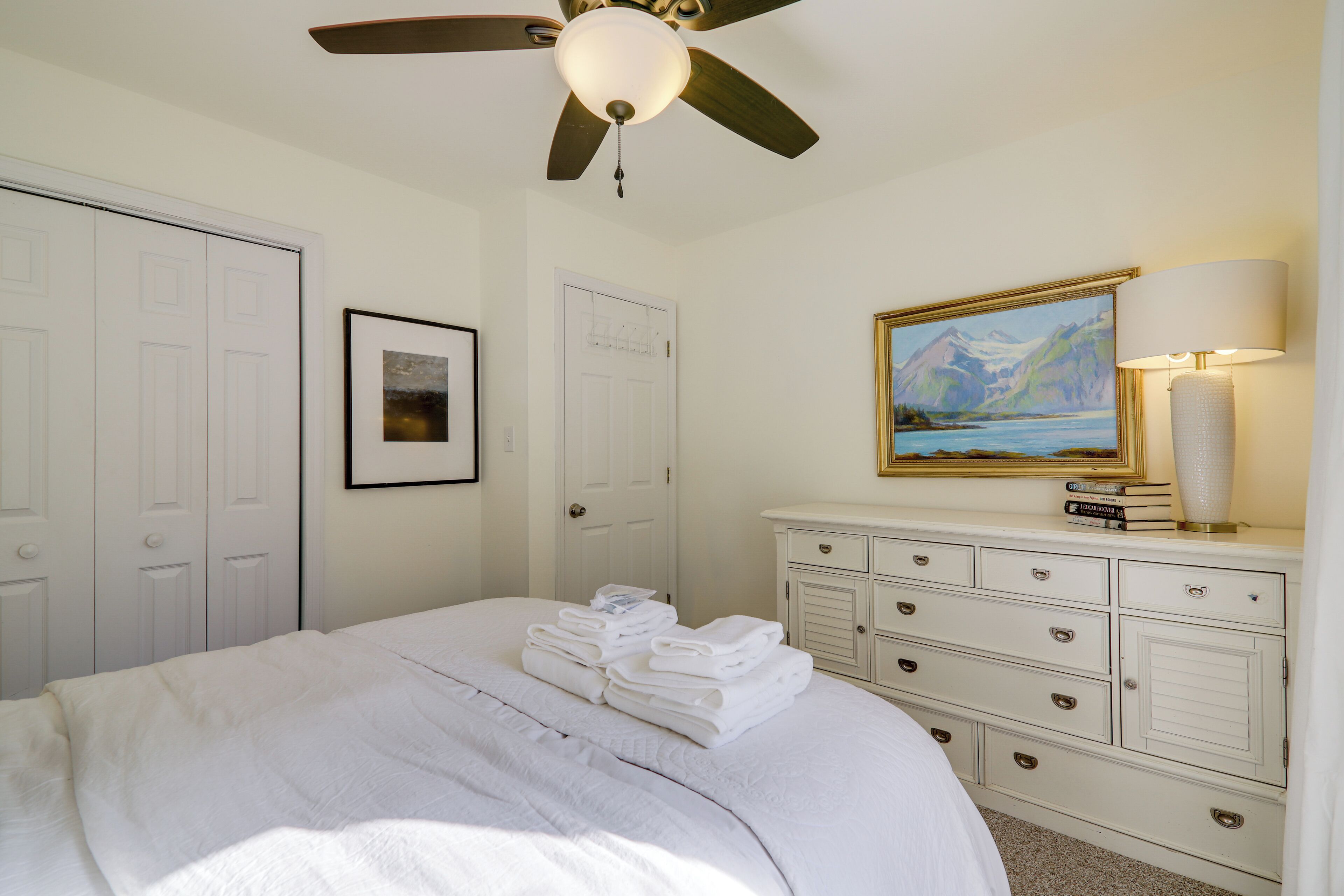 A hotel room with a bed, a ceiling fan, a dresser with drawers, a framed painting, and a lamp.