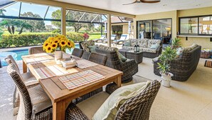 Ein überdachter Außenbereich mit Korbmöbeln, einem Holztisch und Blick auf Pool und Garten.
