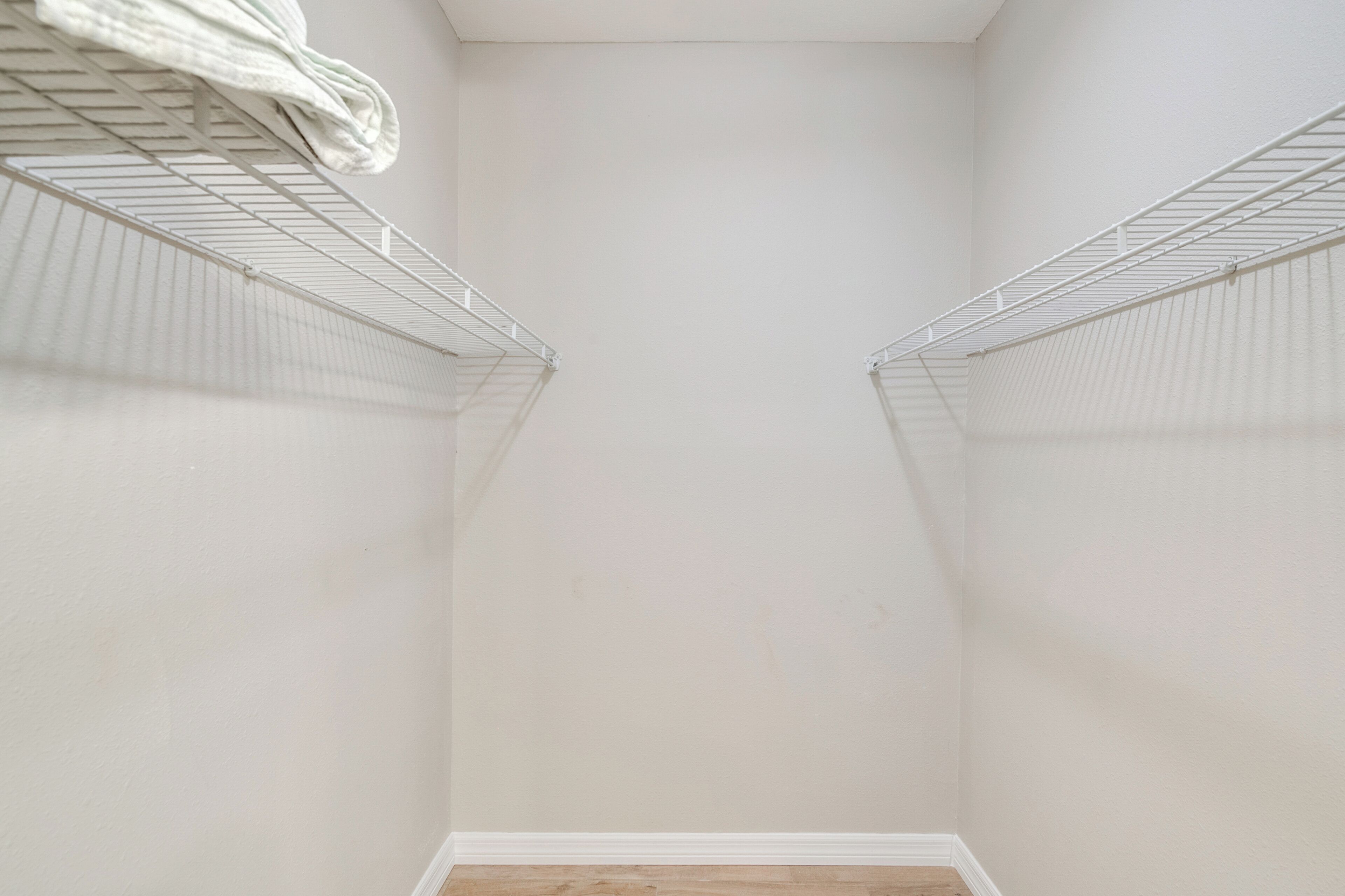 Un placard aux murs blancs et au plancher en bois, doté de deux étagères en fil métallique et d’une tringle pour suspendre les vêtements.