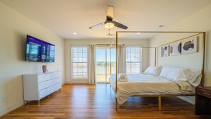 Une chambre à coucher avec un lit, un téléviseur fixé au mur, un ventilateur de plafond et une vue sur l’extérieur, visible à travers la porte.