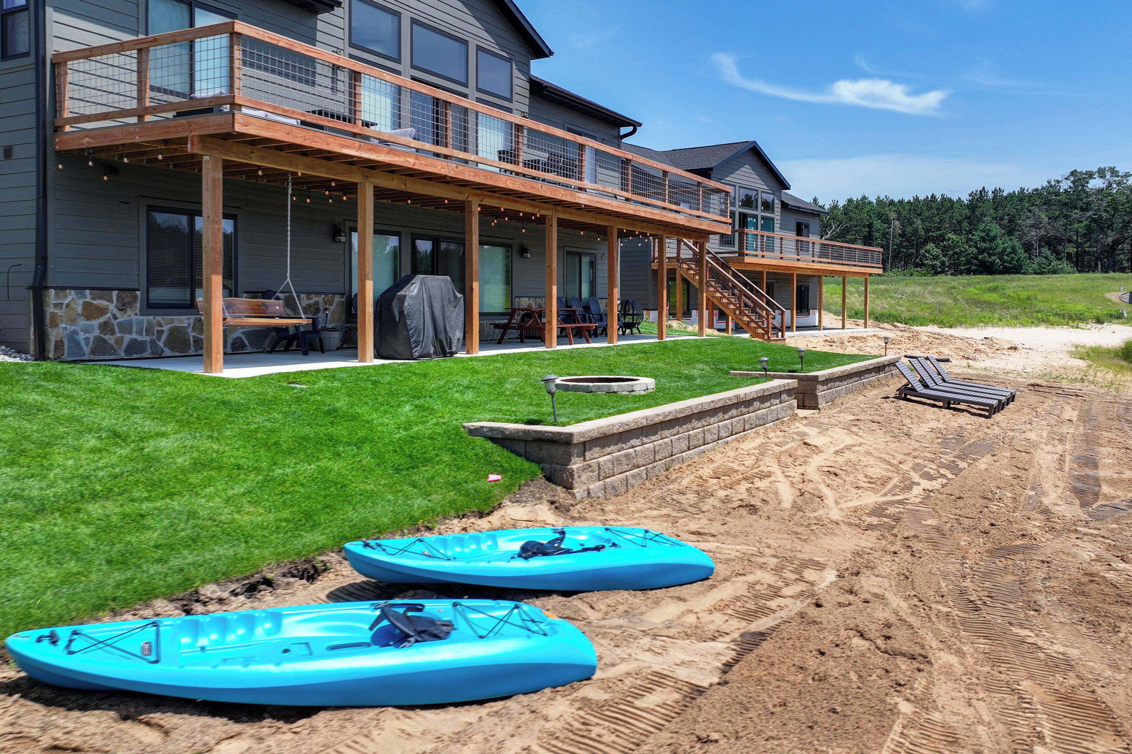 Ein zweistöckiges Haus mit überdachter Terrasse, Sitzgelegenheiten im Freien und einem sandigen Bereich mit Kajaks.