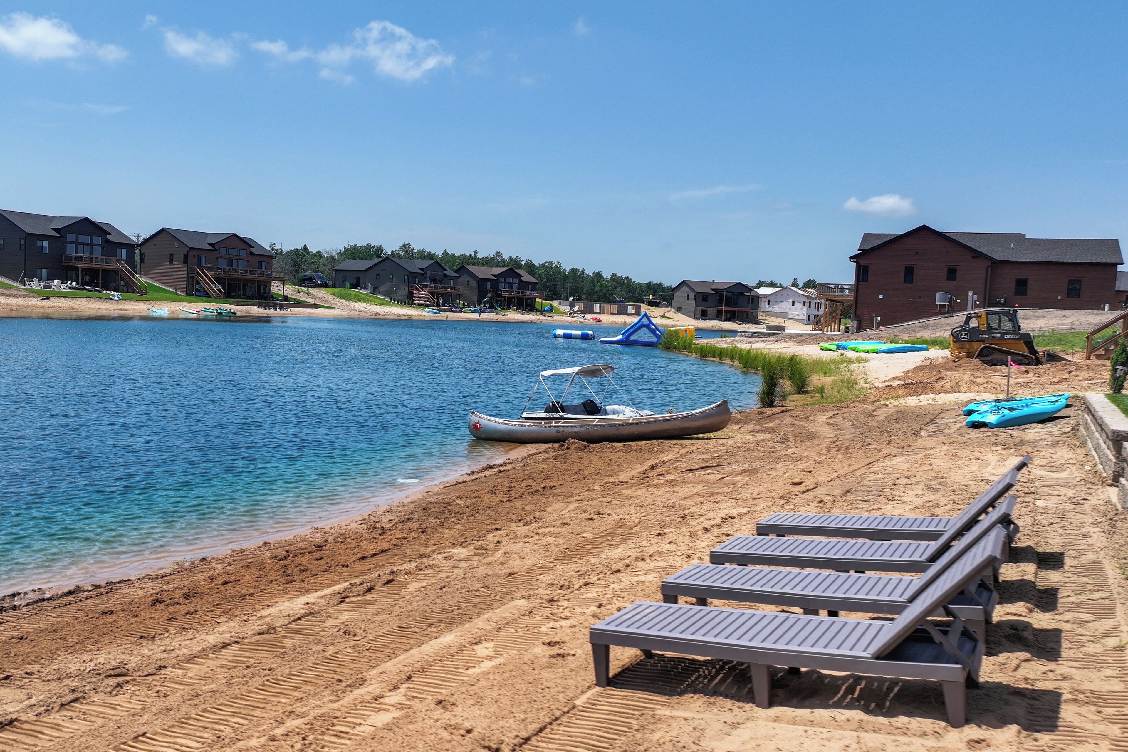 A lakeside property with a sandy beach, lounge chairs, a dock with boats, and a large building.
