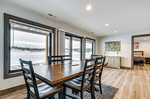 Un comedor con mesa y sillas de madera, ventanas amplias con vistas a un paisaje nevado, y un cuadro en la pared.