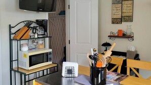 Een keuken met een magnetron, koffiezetapparaat en keukengerei op een plank. Er is een deur, een plank met decoratieve items, en een tafel met stoelen.