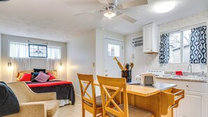 Een kleine kamer met een bed, een eettafel met stoelen, een plafondventilator, een raam met gordijnen en een kitchenette met een spoelbak en aanrecht.
