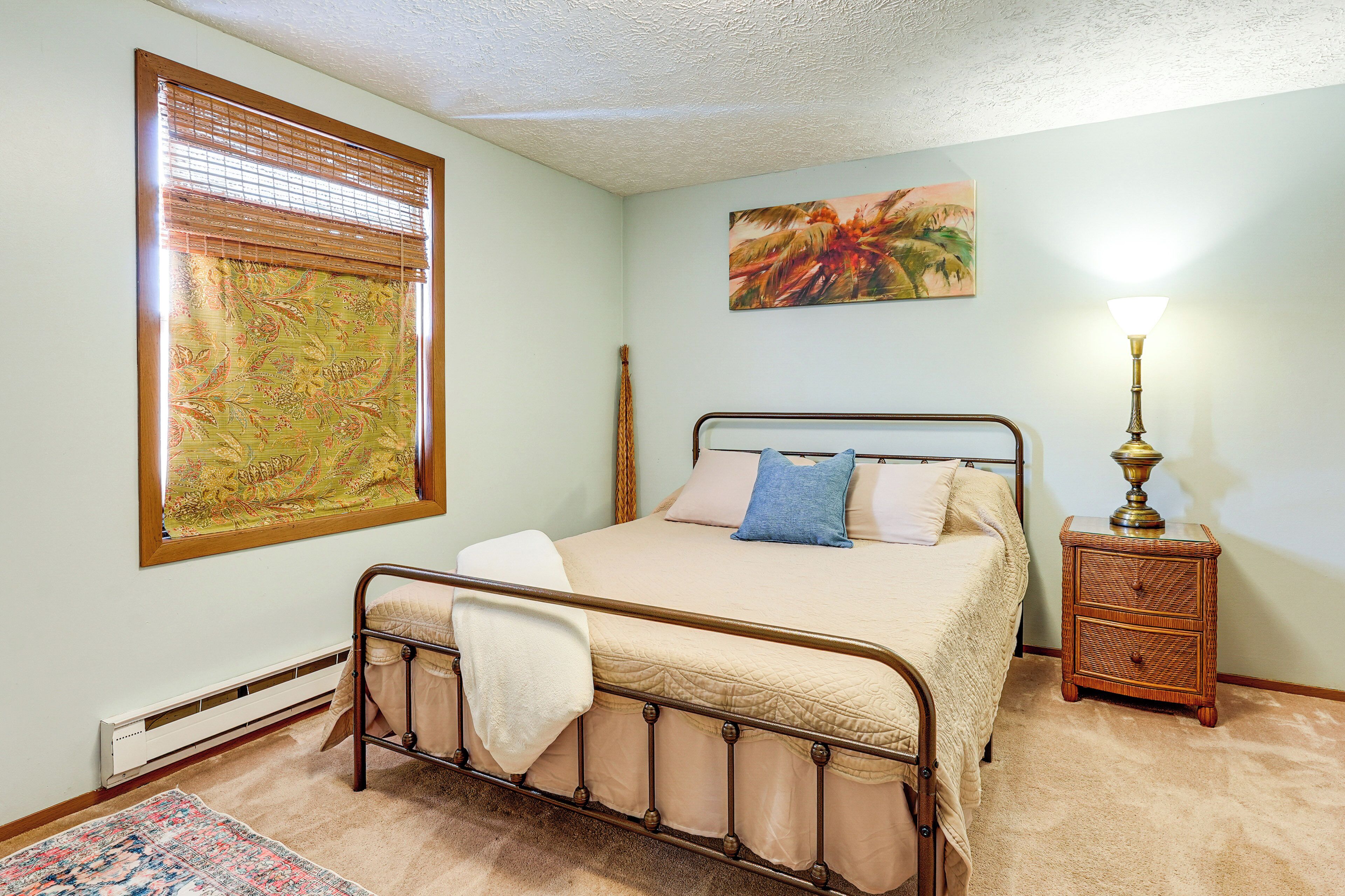 A bedroom with a bed, a nightstand, a lamp, a window with blinds, and a painting on the wall.