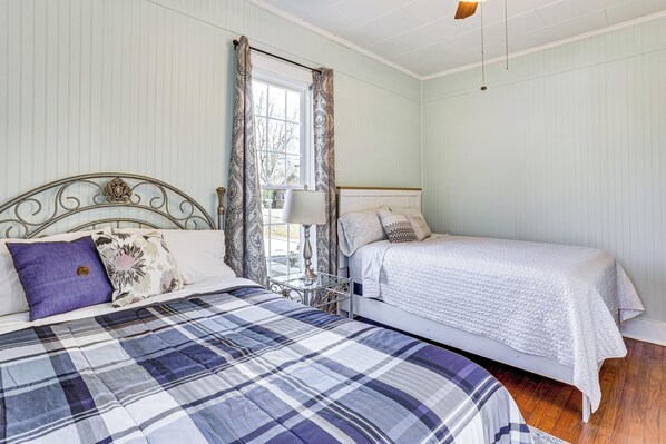 Une chambre avec deux lits, une fenêtre avec des rideaux, une table de chevet avec une lampe et un ventilateur de plafond.
