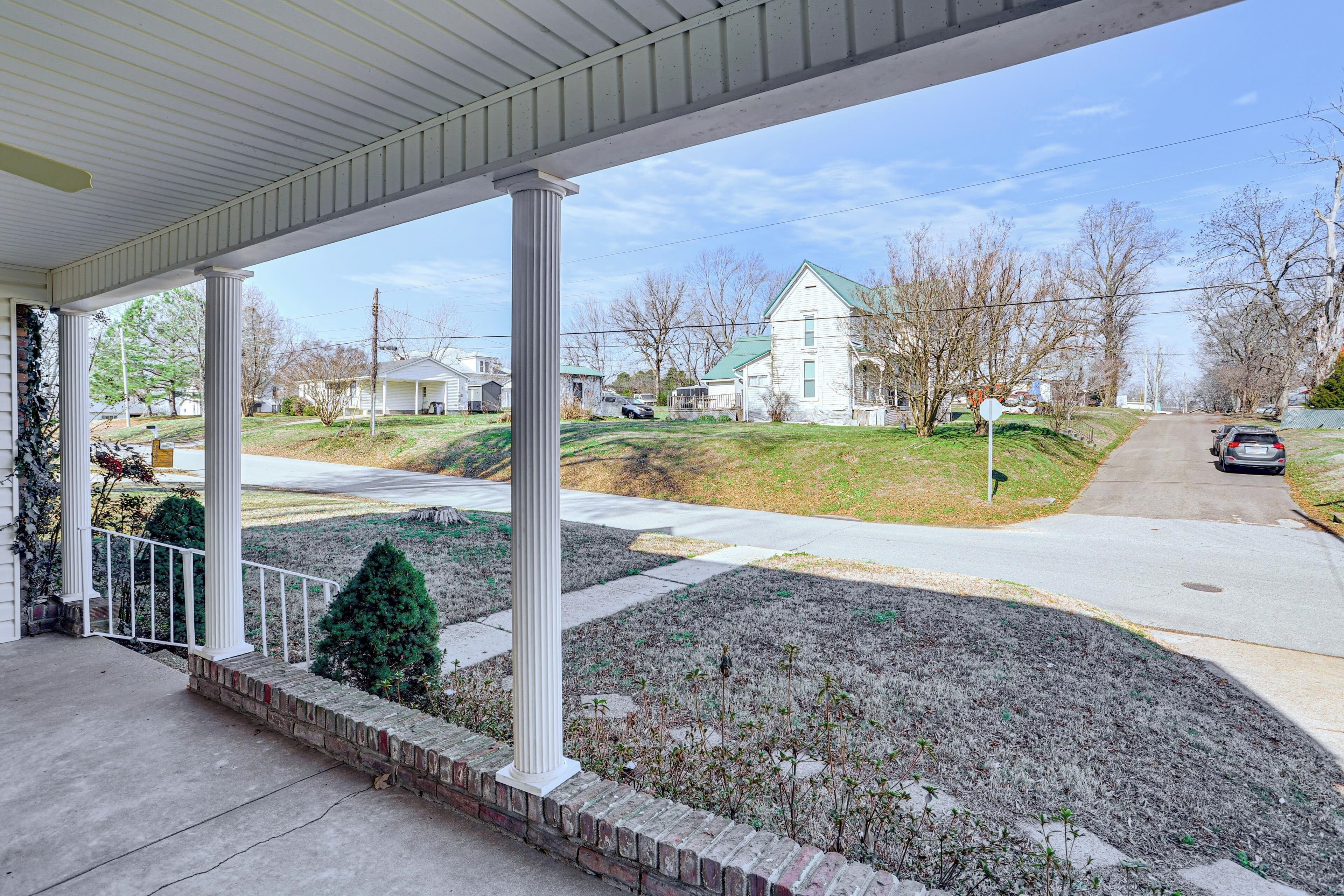 A covered porch with a brick base and white railing, overlooking a residential street with houses and a clear sky.