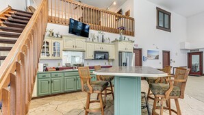 A spacious kitchen with green cabinets, a large island with bar stools, a wooden staircase, and a TV mounted above the island. - Woodburn Group Getaway: Endless On-site Fun! (Harlan)