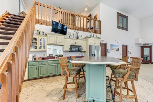 A spacious kitchen with green cabinets, a large island with bar stools, a wooden staircase, and a TV mounted on the wall. - Woodburn Group Getaway: Endless On-site Fun! (Harlan)