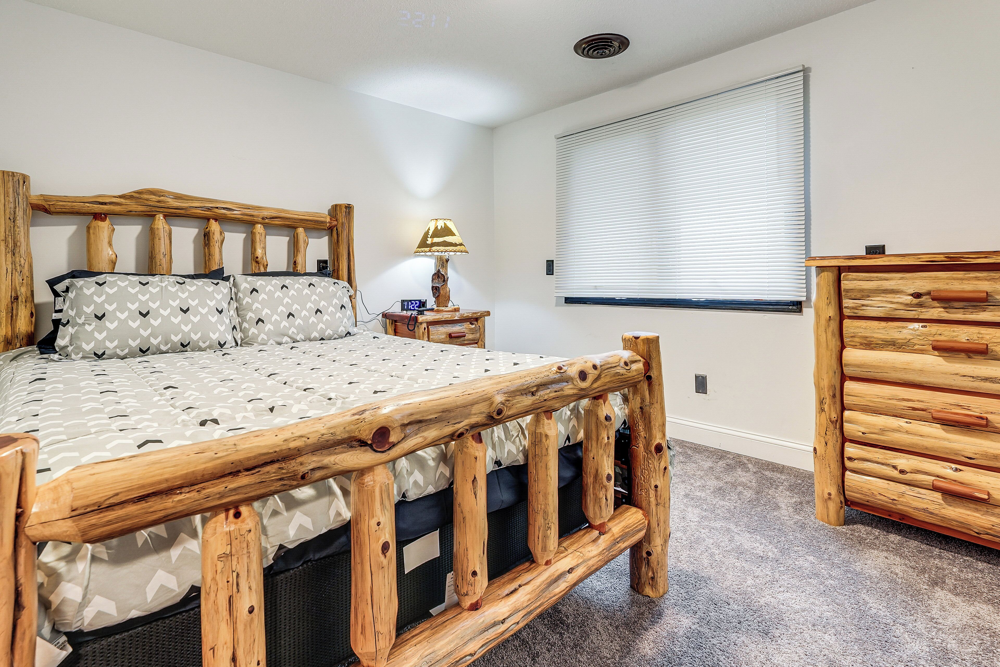 A bedroom with a wooden bed frame, a bedside lamp, a dresser, and a window with blinds.
