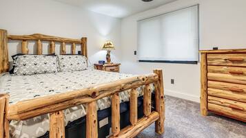 A bedroom with a wooden bed frame, a bedside lamp, a dresser, and a window with blinds.