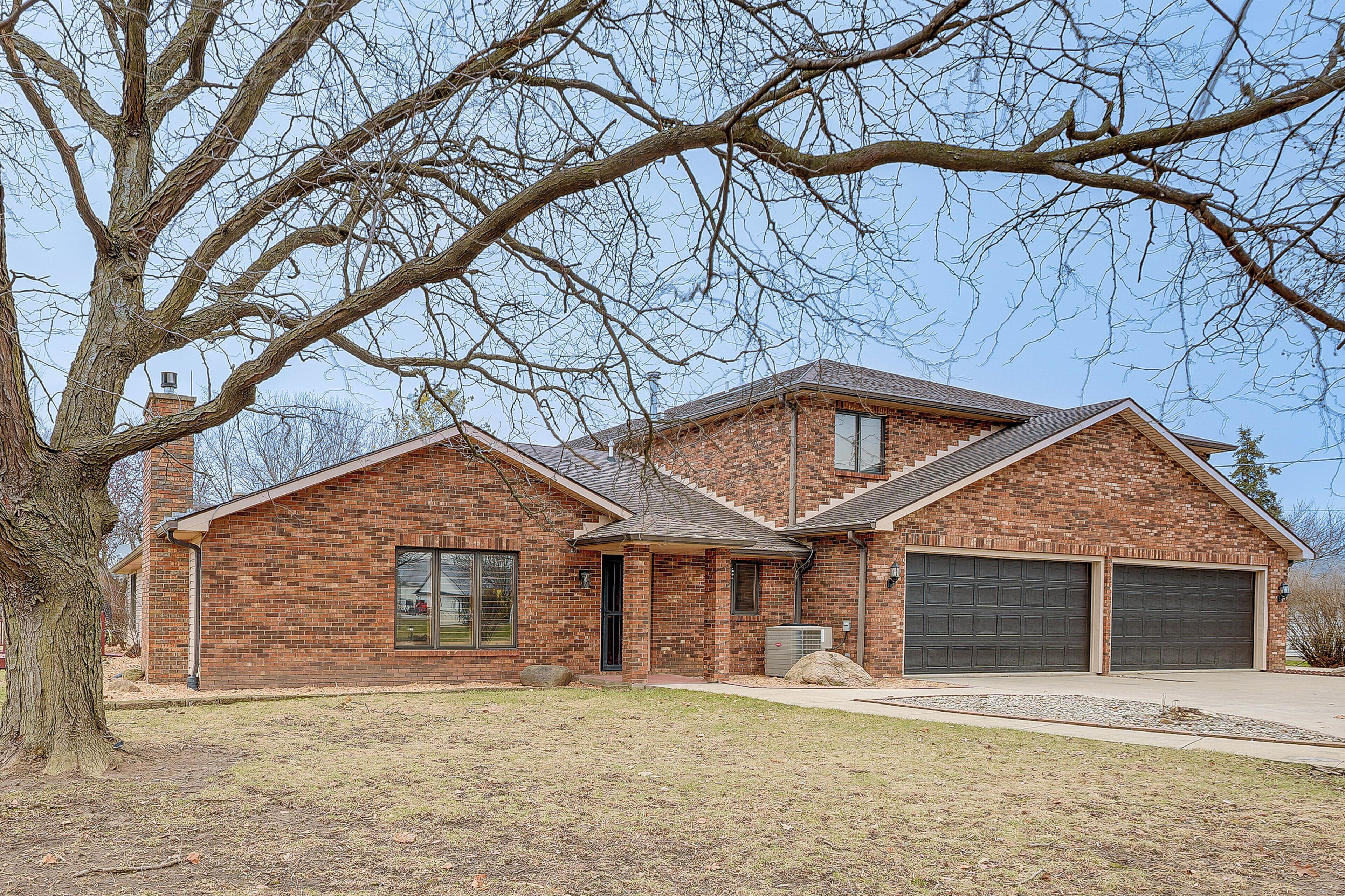 A two-story brick house with a garage, a front porch, and a large tree in the foreground.