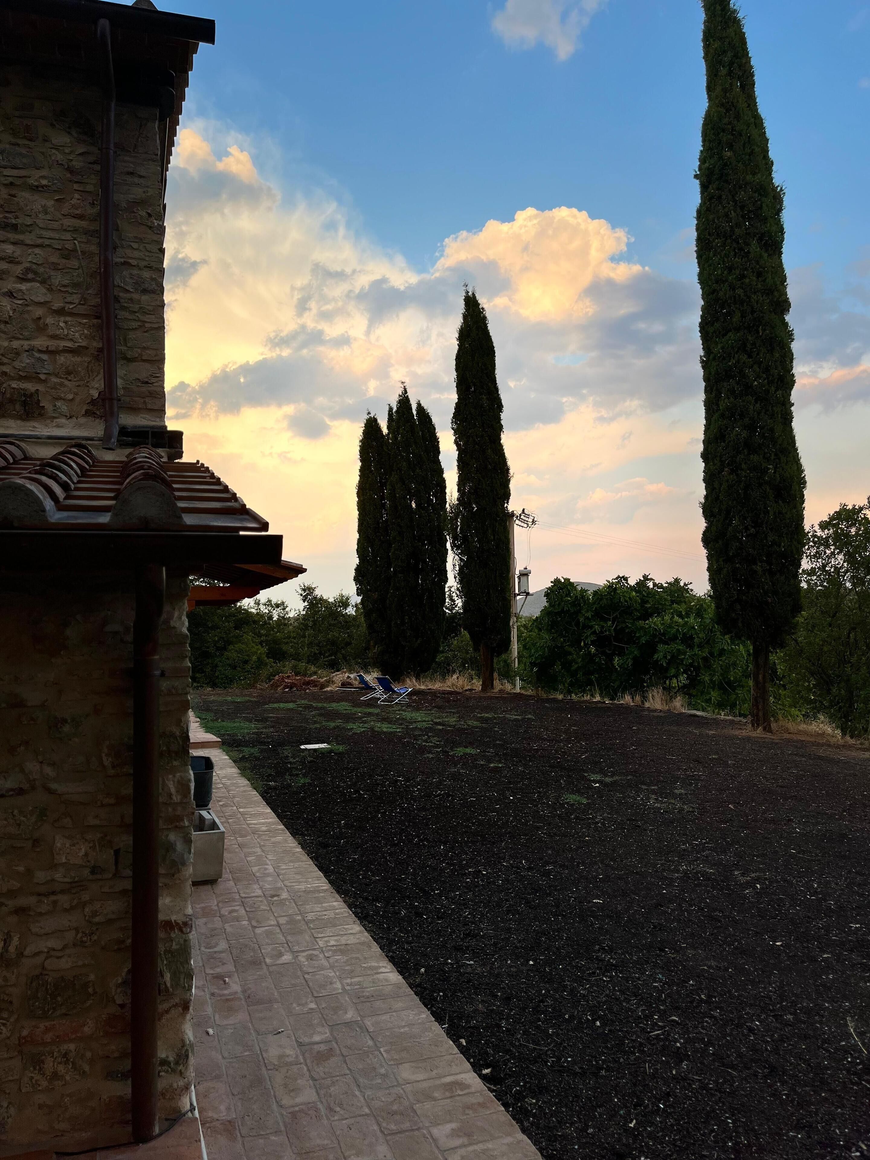 Ein Gebäude aus Stein mit einem Ziegeldach und einem gepflasterten Weg, der zu einer begrünten Anlage mit Bäumen und blauem Himmel führt.