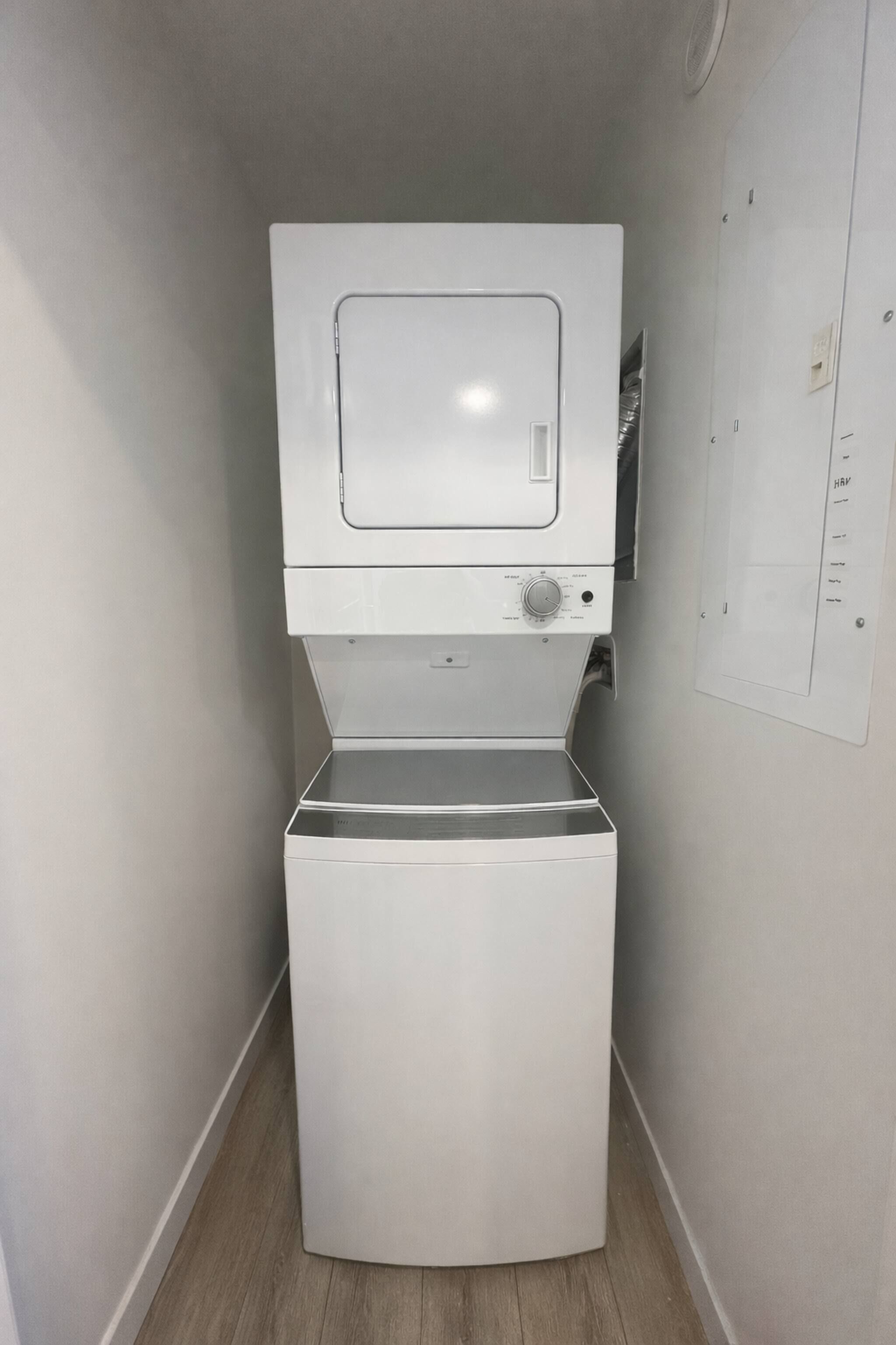 Un lavadero pequeño y blanco con lavadora y secadora integradas, un panel de control y un cuadro eléctrico instalado en la pared.