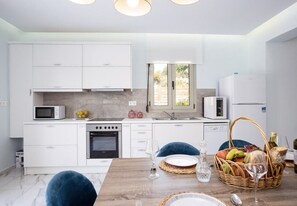 A modern kitchen with white cabinets, a microwave, a stove, and a sink. There is a dining table with chairs and a basket of fruit. - Primavera Villas - 2 Heated Pools Jacuzzis (Rethymno)