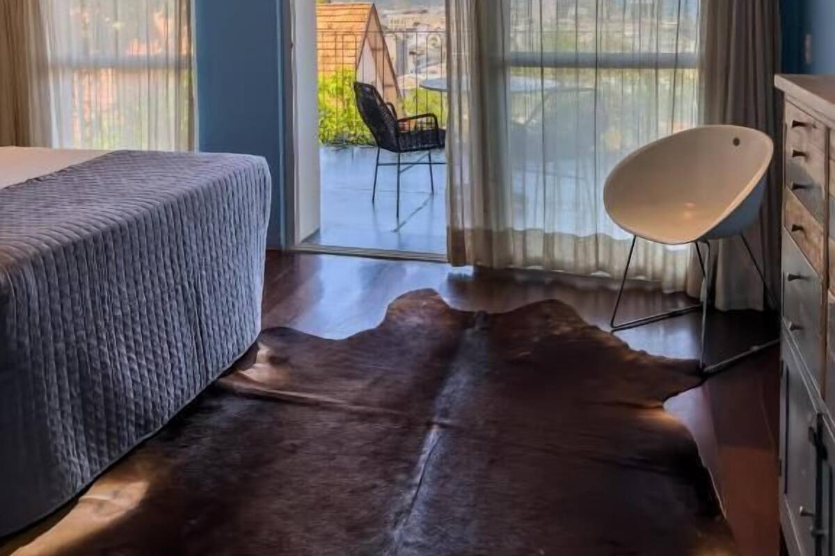 A bedroom with a bed, a chair, a dresser, and a view of the outdoors through large windows.