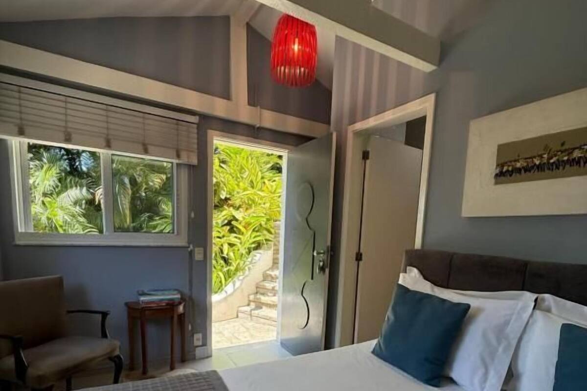 A bedroom with a large bed, a chair, a small table, and a view of greenery outside.
