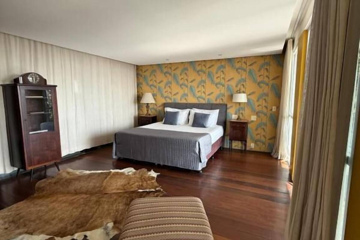 A hotel room with a bed, a wooden dresser, a patterned rug, and a large window with curtains.