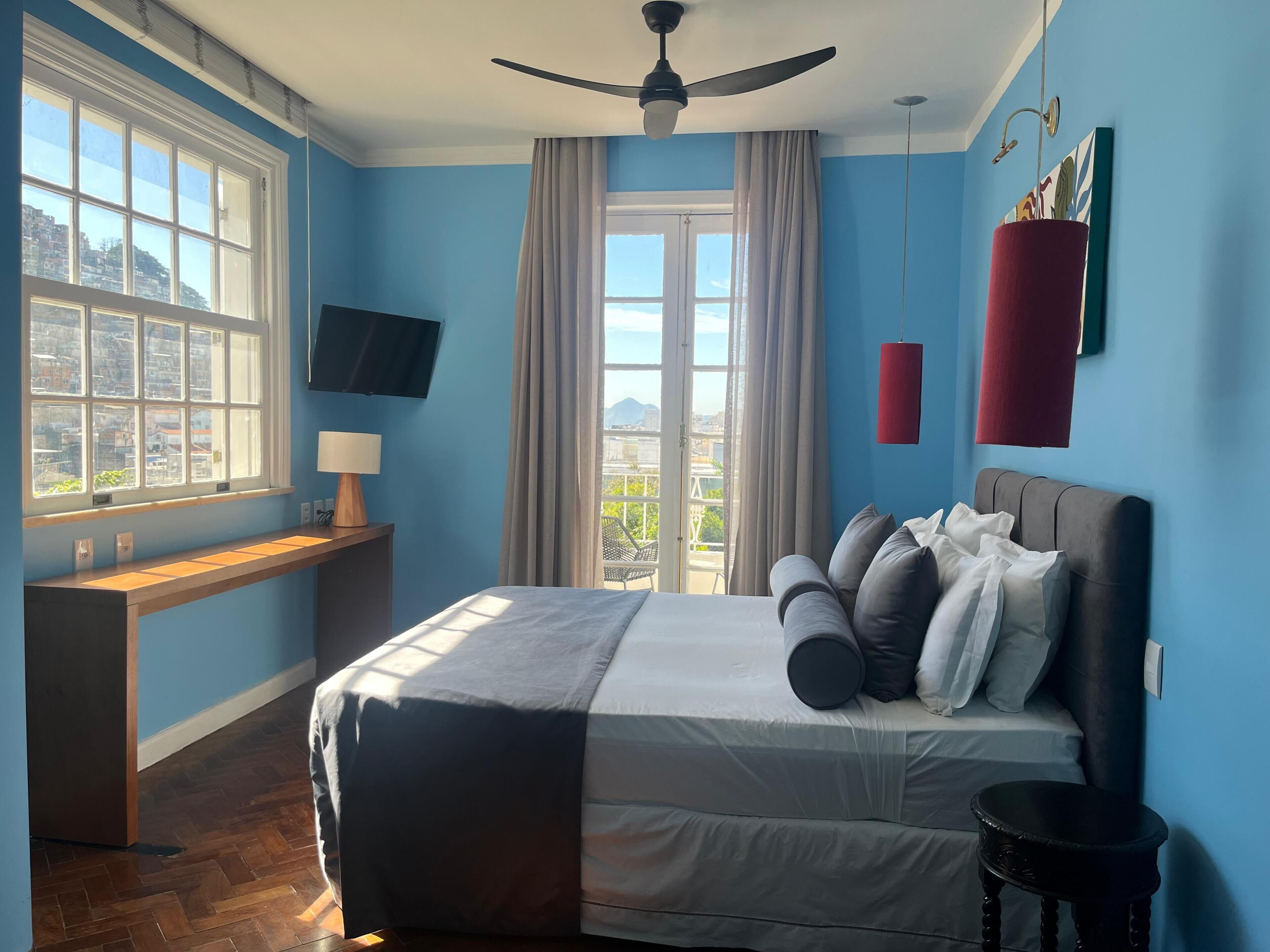 A bedroom with a bed, a desk, a television, a ceiling fan, and a window with curtains.