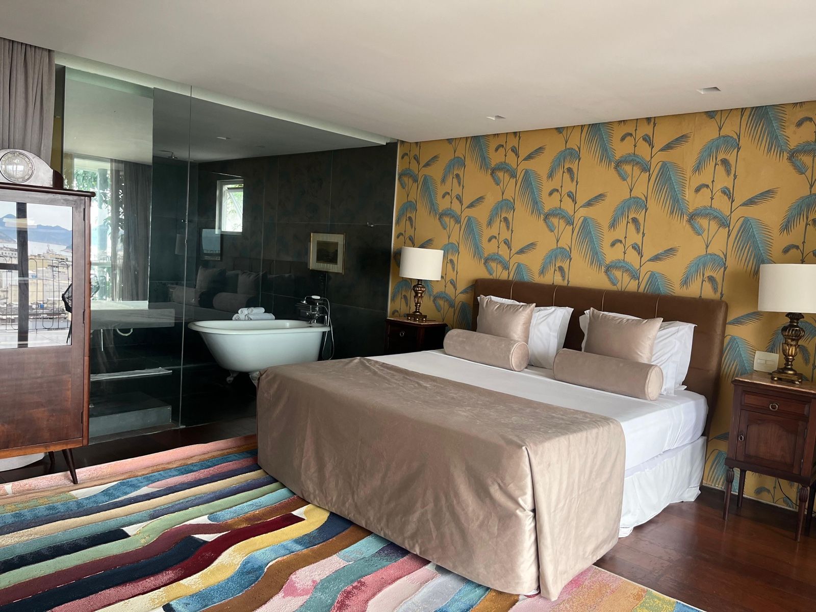 A hotel room with a large bed, a bathtub, a wooden nightstand, and a colorful striped rug.
