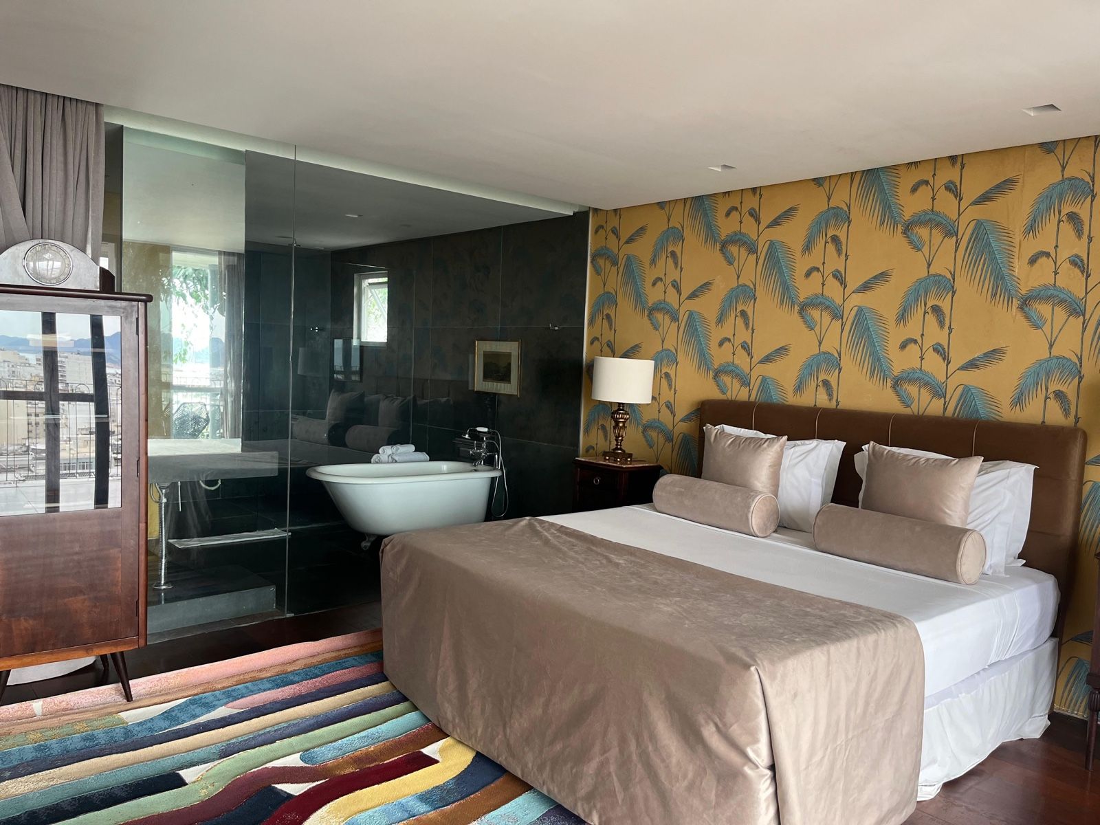 A hotel room with a large bed, a bathtub, a bedside table with a lamp, and a colorful rug.