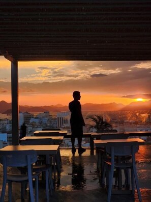 A person standing at a table with a view of a sunset over a cityscape.
