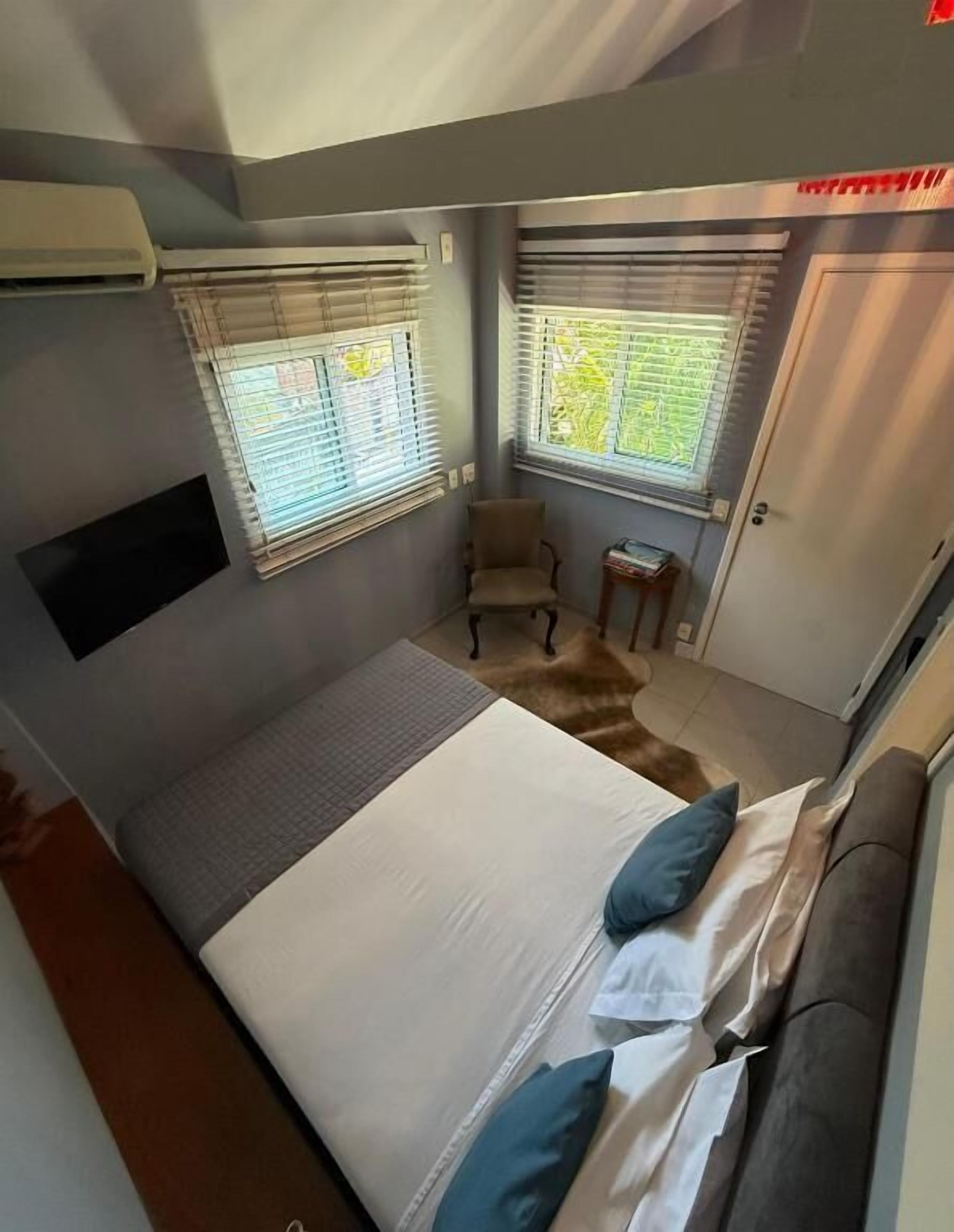 A bedroom with a bed, two windows with blinds, a chair, a small table, and a door.