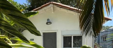 Un pequeño edificio de una planta con fachada blanca y tejado rojo. Tiene una puerta y una ventana con contraventanas, rodeado de abundante vegetación verde.