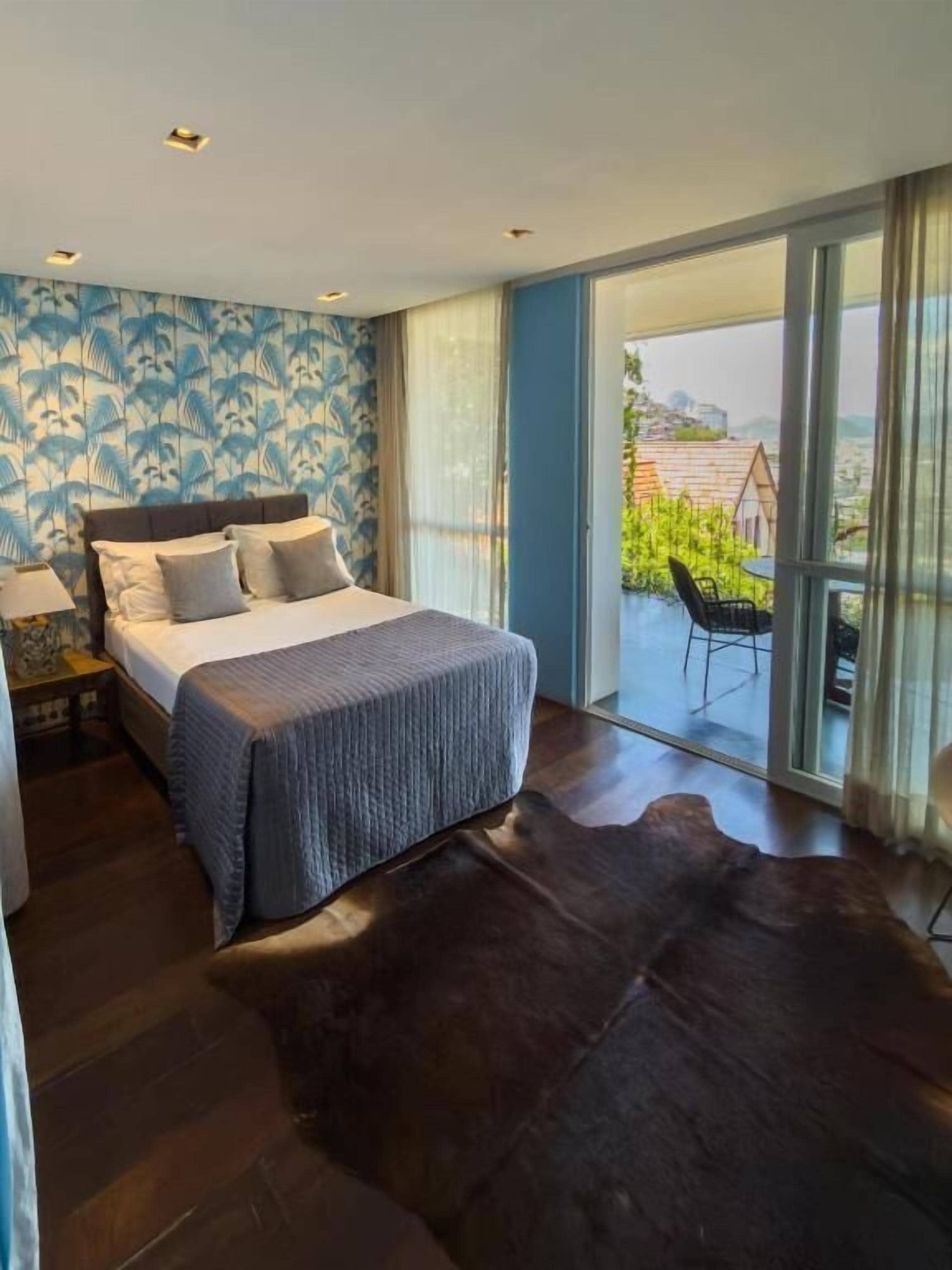 A bedroom with a large bed, a nightstand, a window with a view, and a balcony with a chair.