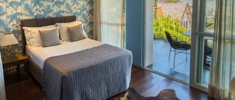 A bedroom with a large bed, a nightstand, a window with a view, and a balcony with a chair.