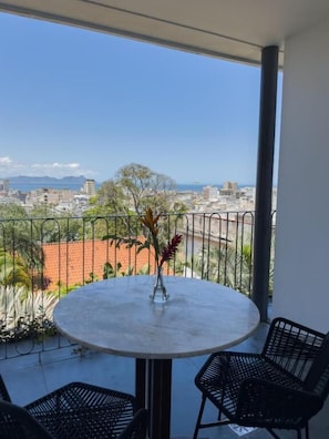 Ein Balkon mit rundem Tisch und zwei Stühlen, mit Blick auf eine Stadtlandschaft mit Gebäuden und einer entfernten Bergsicht.