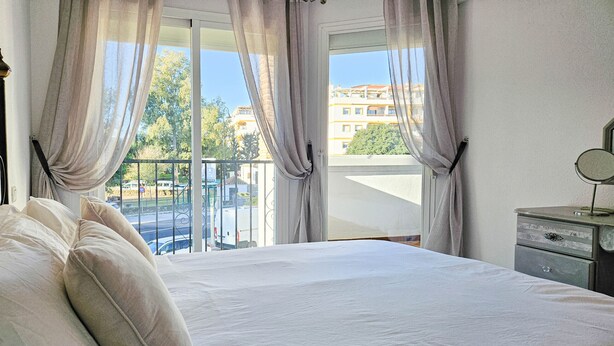 Une chambre avec un grand lit, une table de chevet avec un miroir, et une vue sur un immeuble et une rue, visible par la fenêtre.