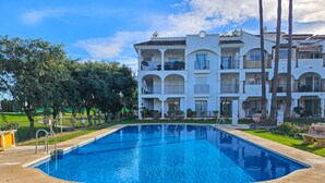 Une piscine entourée d’une pelouse bien entretenue, avec un immeuble à plusieurs étages doté de balcons et d’un toit en tuiles en arrière-plan.