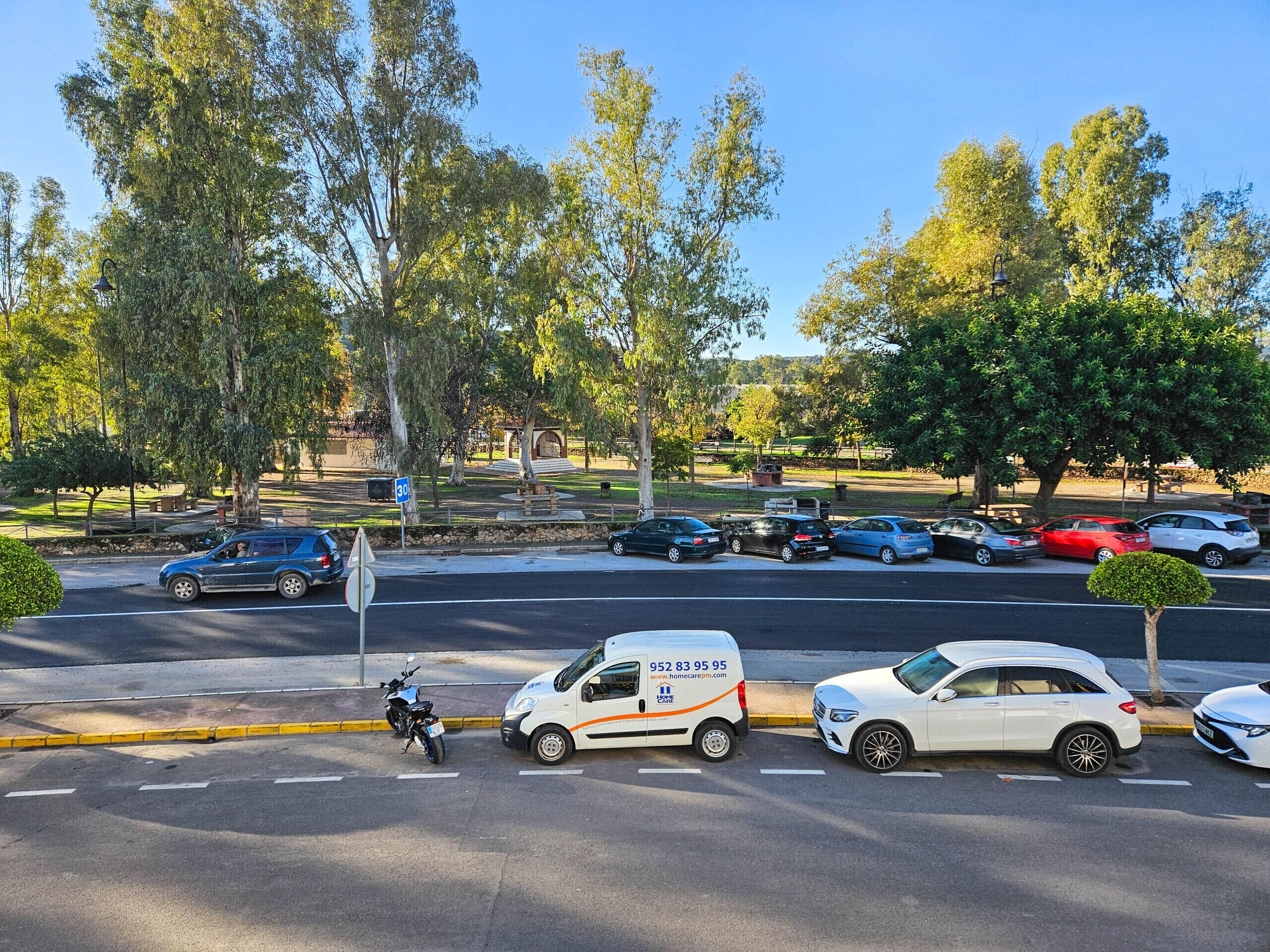 Une vue d’une rue avec des voitures garées, une moto et un fourgon sur lequel figure un numéro de téléphone.