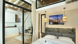 Een slaapkamer met een houten hoofdeinde, een bed met witte lakens, een schilderij aan de muur en een trap naar een balkon.